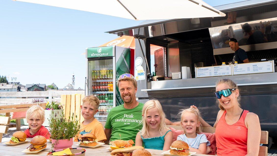 Eine Gruppe von fünf Personen sitzt an einem Tisch im Freien und genießt große Hamburger. Im Hintergrund ist ein Imbisswagen und eine angenehme Sommeratmosphäre zu sehen. | © Henrieke Ibing