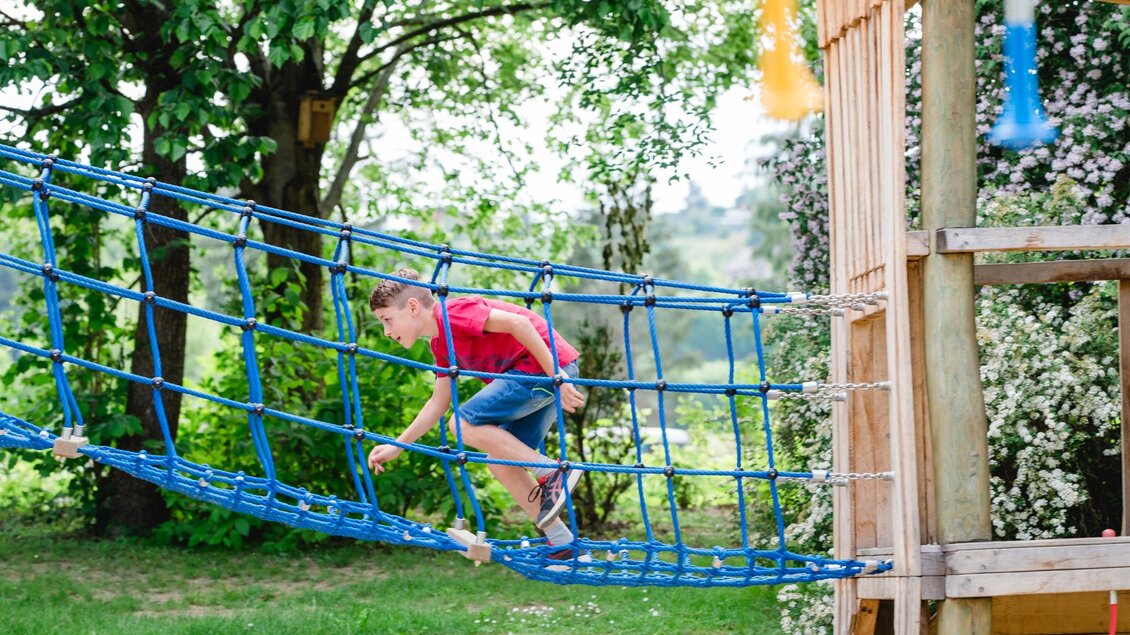 Ein Junge spielt auf einem blauen Klettergerüst in einer grünen Gartenlandschaft. Im Hintergrund sind Bäume und blühende Sträucher zu sehen. | © Miasphotoart