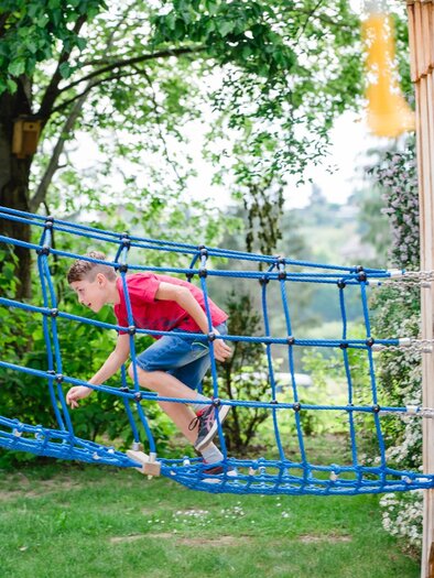 A boy is playing on a blue climbing frame in a green garden landscape. In the background, trees and blooming bushes can be seen. | © Miasphotoart