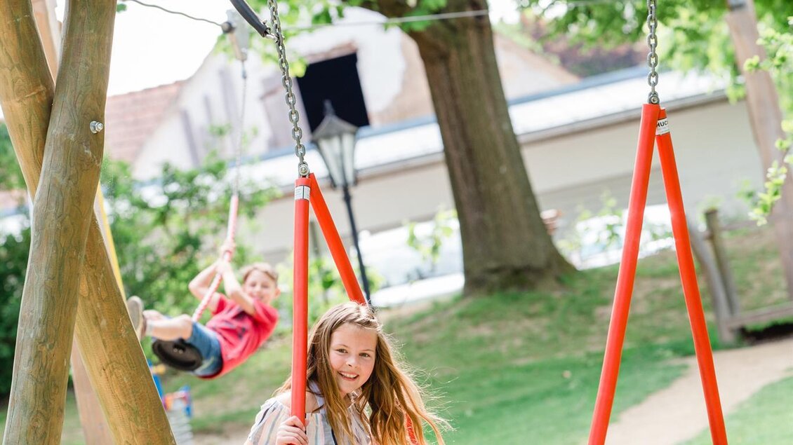 Ein Spielplatz mit einer großen Nestschaukel, auf der ein Mädchen sitzt. Im Hintergrund schwingt ein Junge auf einer anderen Schaukel. | © Miasphotoart