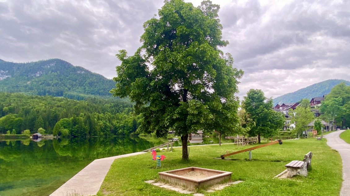 Ein ruhiger See mit grünen Wiesen und Bäumen. Im Hintergrund sind Berge und ein paar Gebäude zu sehen. | © Bettina Scheck