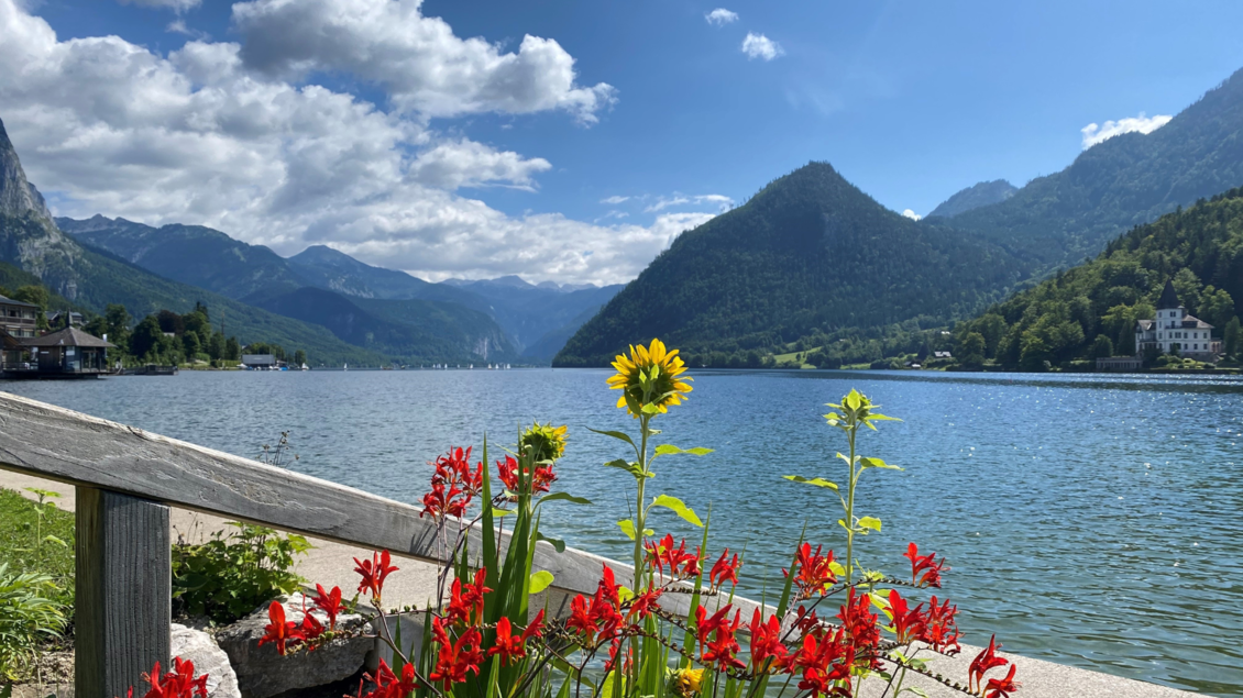 Ein ruhiger See, umgeben von hohen Bergen und üppigem Grün. Im Vordergrund blühen bunte Blumen, die dem Bild Leben verleihen. | © Bettina Scheck