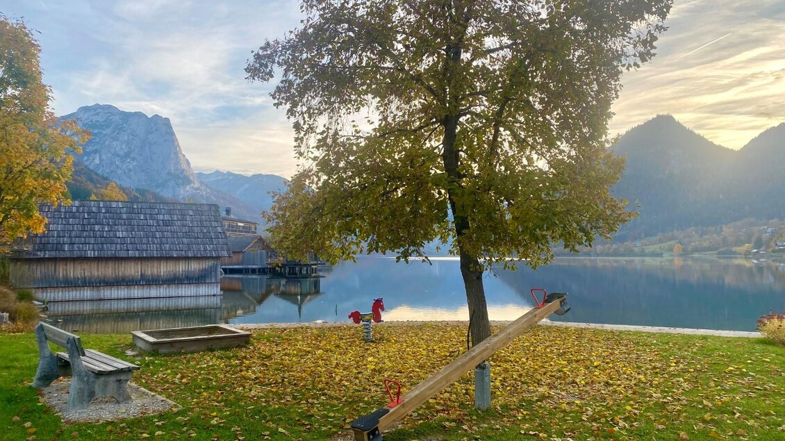 Ein ruhiger See mit einem Baum und einer Schaukel im Vordergrund. Im Hintergrund sind Berge und ein Holzgebäude zu sehen. | © Bettina Scheck