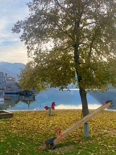 A tranquil lake with a tree and a swing in the foreground. In the background, mountains and a wooden building can be seen. | © Bettina Scheck