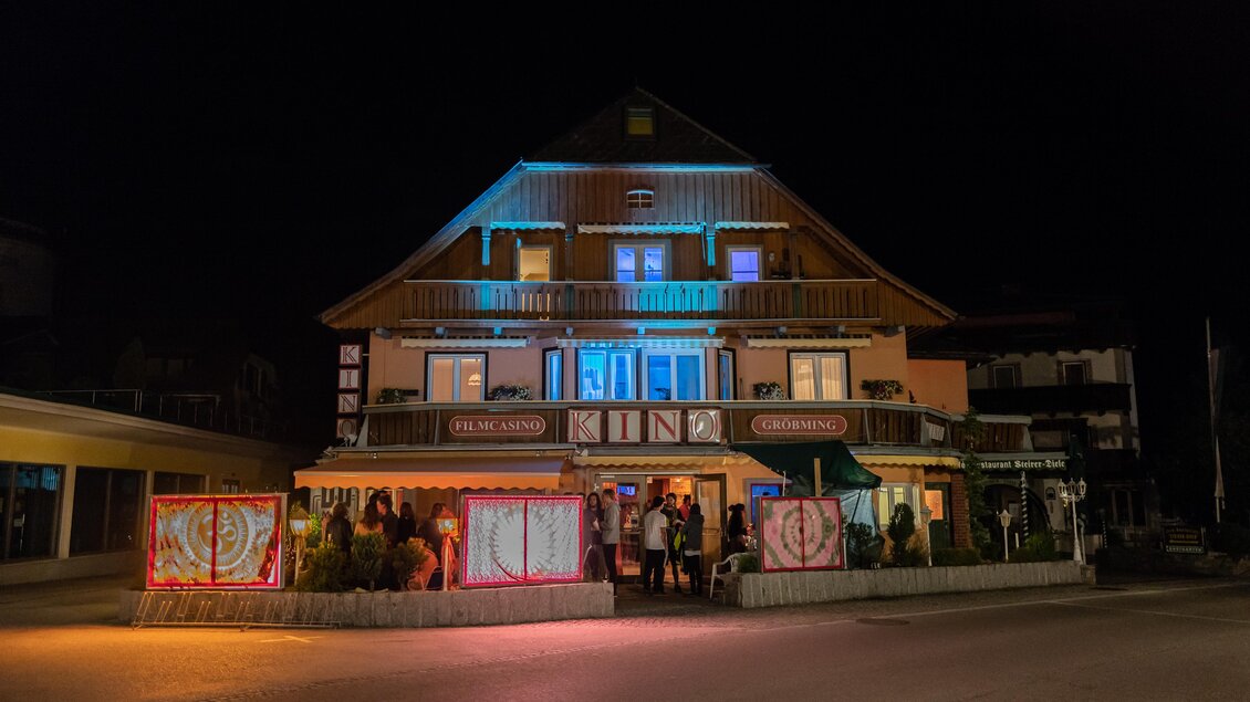 Ein beleuchtetes Gebäude bei Nacht mit einem Restaurant und einer einladenden Atmosphäre. Die Fassade hat eine freundliche Ausstrahlung und zieht Passanten an. | © Kino Gröbming