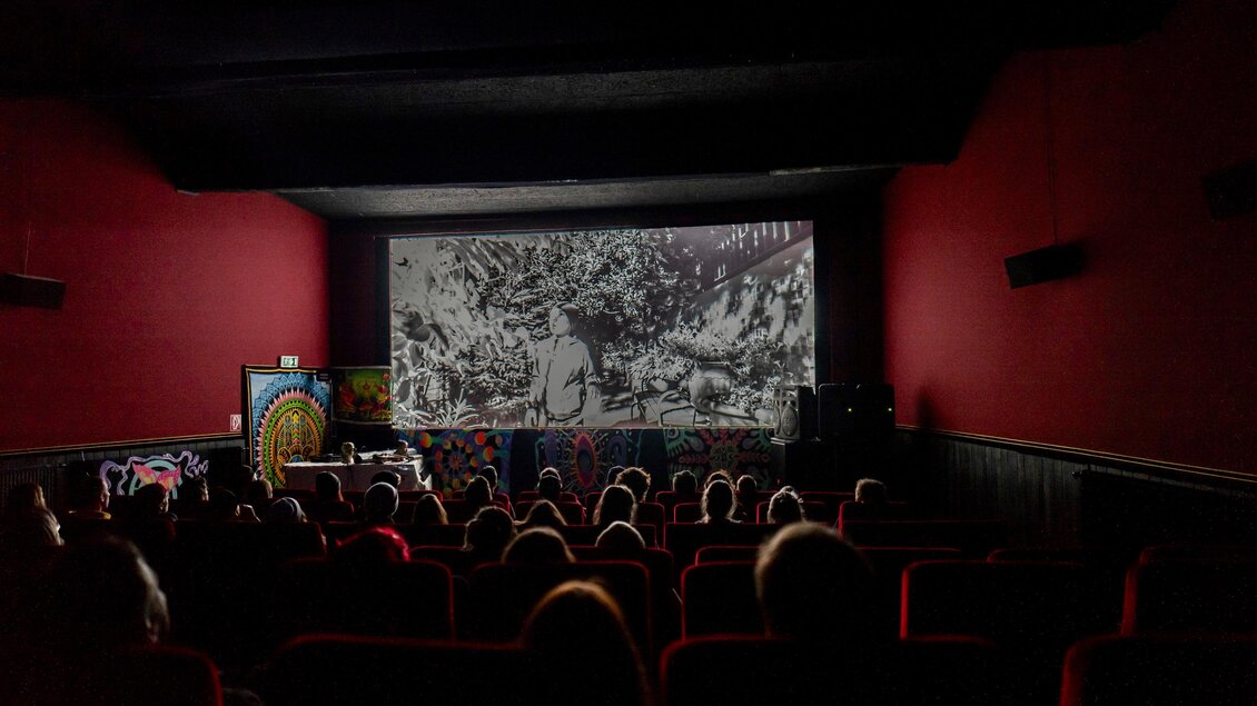 Ein Kino mit roten Wänden und vielen Zuschauern, die auf eine große Leinwand schauen. Auf der Leinwand läuft ein Schwarz-Weiß-Film. | © Kino Gröbming