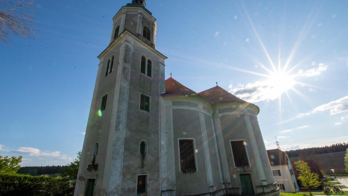 Eine alte Kirche mit einem hohen Turm und einem grünen Garten. Die Sonne scheint hell hinter dem Gebäude. | © Kurkommission Bad Blumau
