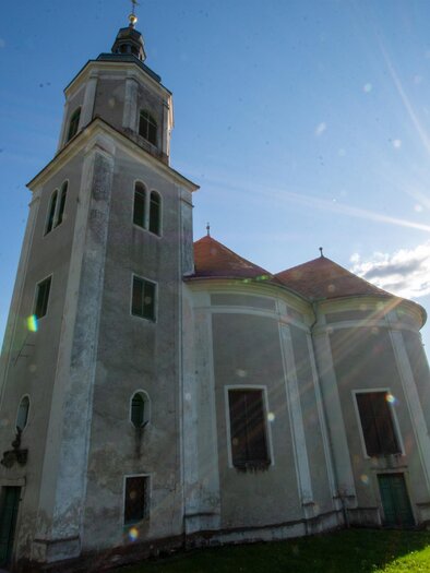 Eine alte Kirche mit einem hohen Turm und einem grünen Garten. Die Sonne scheint hell hinter dem Gebäude. | © Kurkommission Bad Blumau