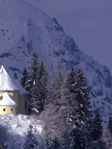 Eine malerische Kirche steht auf einem schneebedeckten Hügel, umgeben von hochgewachsenen Tannen. Im Hintergrund erheben sich majestätische Berge. | ©  TVB Ausseerland - Salzkammergut_Pirker