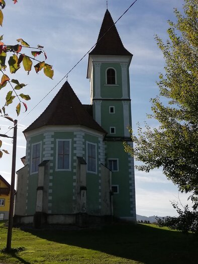 Eine malerische Kirche mit einem grünen Anstrich und einem spitzen Turm, umgeben von Bäumen und Wiese. Die Sonne scheint im Hintergrund und schafft eine idyllische Atmosphäre.