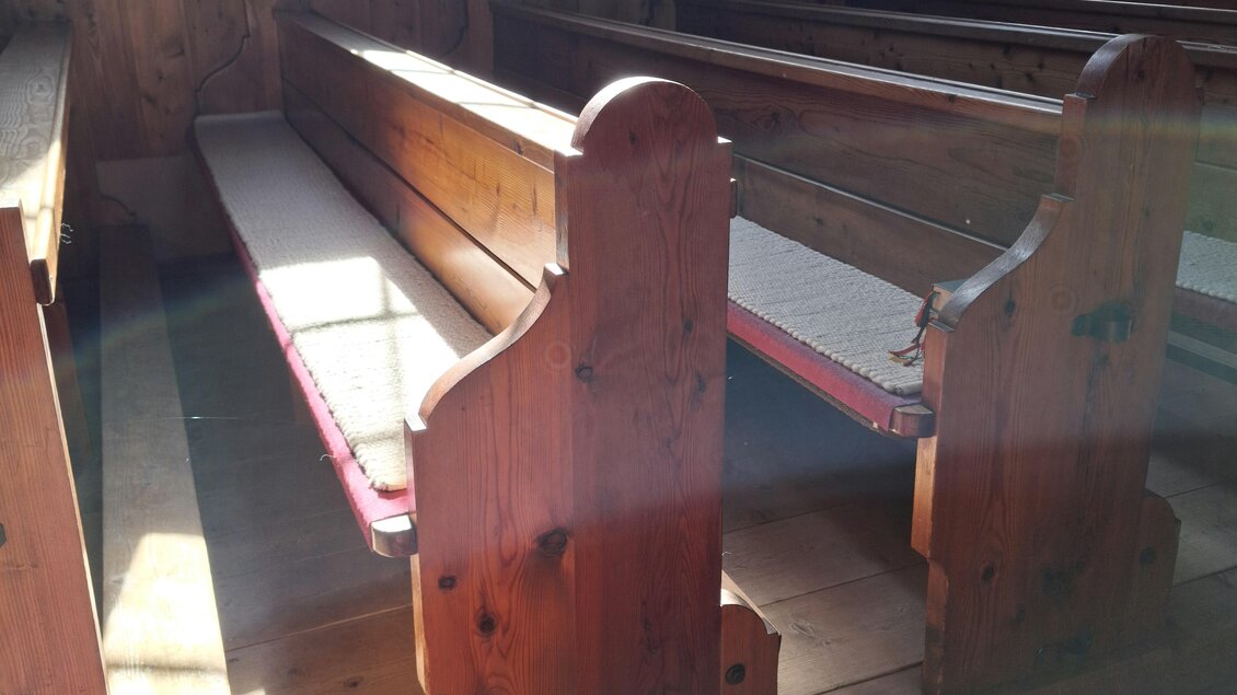 Ein Innenraum mit hölzernen Bänken in einer Kirche. Das Licht fällt sanft auf die Bänke und erzeugt eine ruhige Atmosphäre. | © Beate Koller