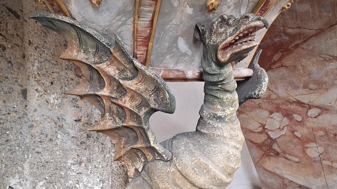 Ein dekorativer Drache mit Flügeln aus Stein steht unter einem kunstvoll verzierten Baldachin. Der Hintergrund zeigt eine strukturierte Wand mit goldenen Elementen. | © Beate Koller
