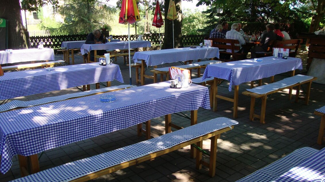 Biergarten mit blauweiß gedeckten Holztischen und Bänken unter schattigen Bäumen, einige Gäste sitzen im Hintergrund. | © Fam. Fauland