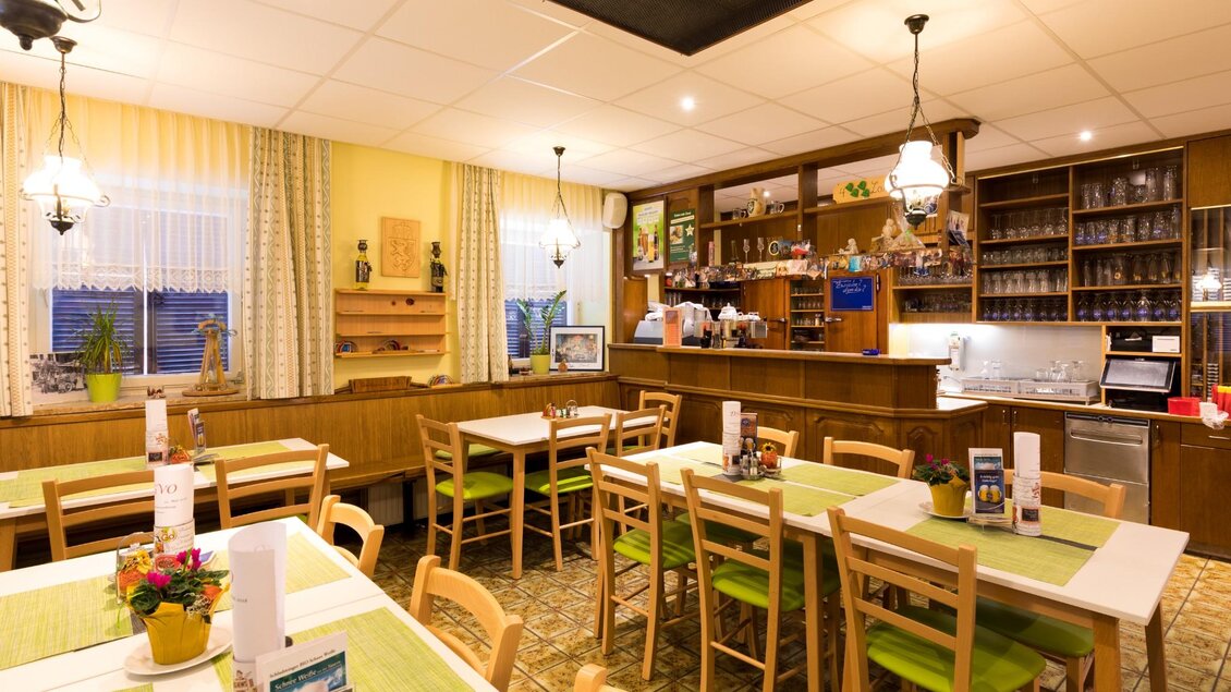 Heller Gastraum mit gedeckten Tischen, grünen Sitzpolstern und einer Holzschank mit Gläsern und Deko im Hintergrund. | © TV Region Graz - Harry Schiffer