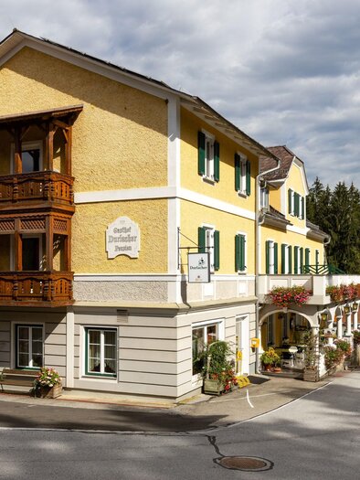 Ein charmantes gelbes Gebäude mit Holzbalustrade und grünen Fenstern. Im Hintergrund ist ein Kirchturm und Bäume zu sehen. | © Gasthof Durlacher