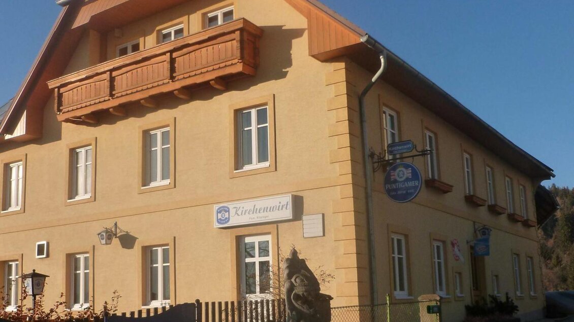 Ein hellgelbes Gebäude mit einem Balkon und mehreren Fenstern. Vor dem Gebäude befindet sich ein Zaun und eine kleine Statue. | © Kirchenwirt Gasthaus Ritzinger