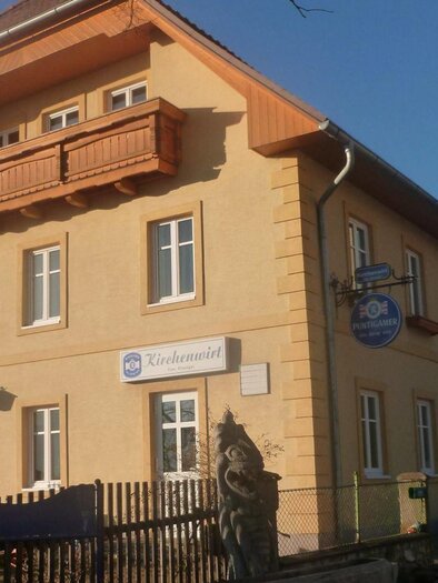 Ein hellgelbes Gebäude mit einem Balkon und mehreren Fenstern. Vor dem Gebäude befindet sich ein Zaun und eine kleine Statue. | © Kirchenwirt Gasthaus Ritzinger