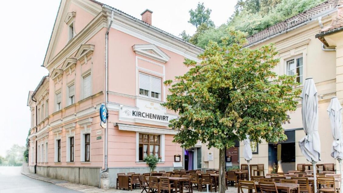 Ein charmantes Restaurant namens "Kirchenwirt" mit einem rosa Außenanstrich. Draußen stehen Tische und Stühle unter einem grünen Baum. | © Kirchenwirt an der Weinstraße | Oliver Haring