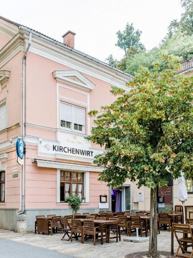 A charming restaurant called "Kirchenwirt" with a pink exterior. Outside, there are tables and chairs under a green tree. | © Kirchenwirt an der Weinstraße | Oliver Haring