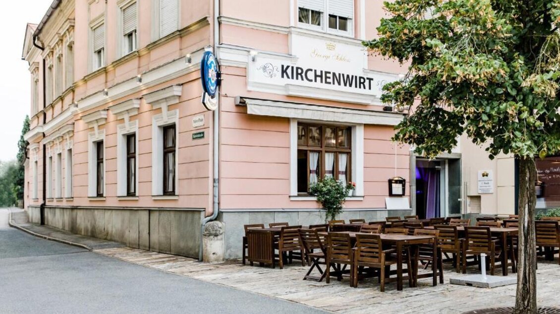 Ein hübsches Restaurant mit dem Namen "Kirchenwirt" an einer ruhigen Straße. Draußen stehen Holz-Tische und -Stühle für Gäste. | © Kirchenwirt an der Weinstraße | Oliver Haring