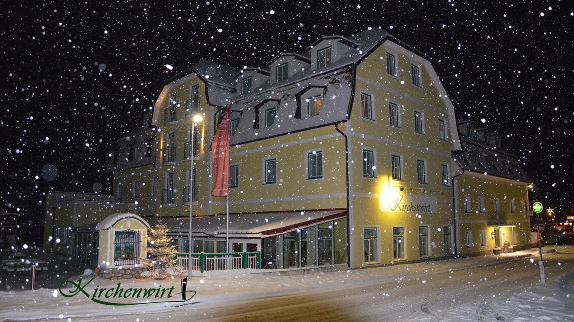 Ein Hotelgebäude in einer verschneiten Landschaft bei Nacht. Schneeflocken fallen sanft auf die Straßen und das Gebäude. | © Kirchenwirt Aigen