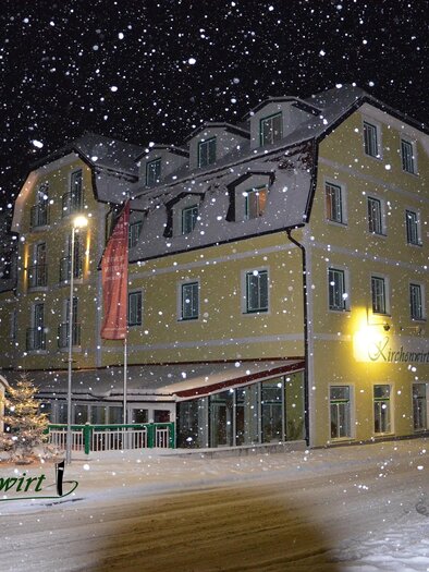 A hotel building in a snowy landscape at night. Snowflakes gently fall on the streets and the building. | © Kirchenwirt Aigen