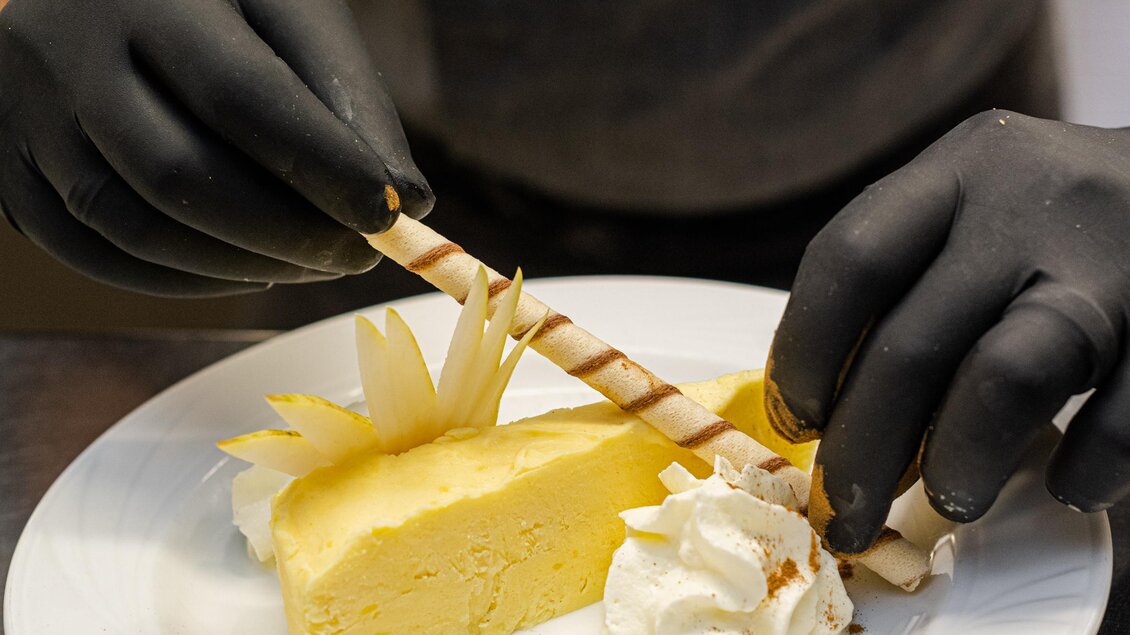 Ein Stück Käsekuchen wird kunstvoll mit Sahne und Dekoration auf einem Teller angerichtet. Der Koch trägt schwarze Handschuhe und konzentriert sich auf die Präsentation. | © Martina Siebenhandl