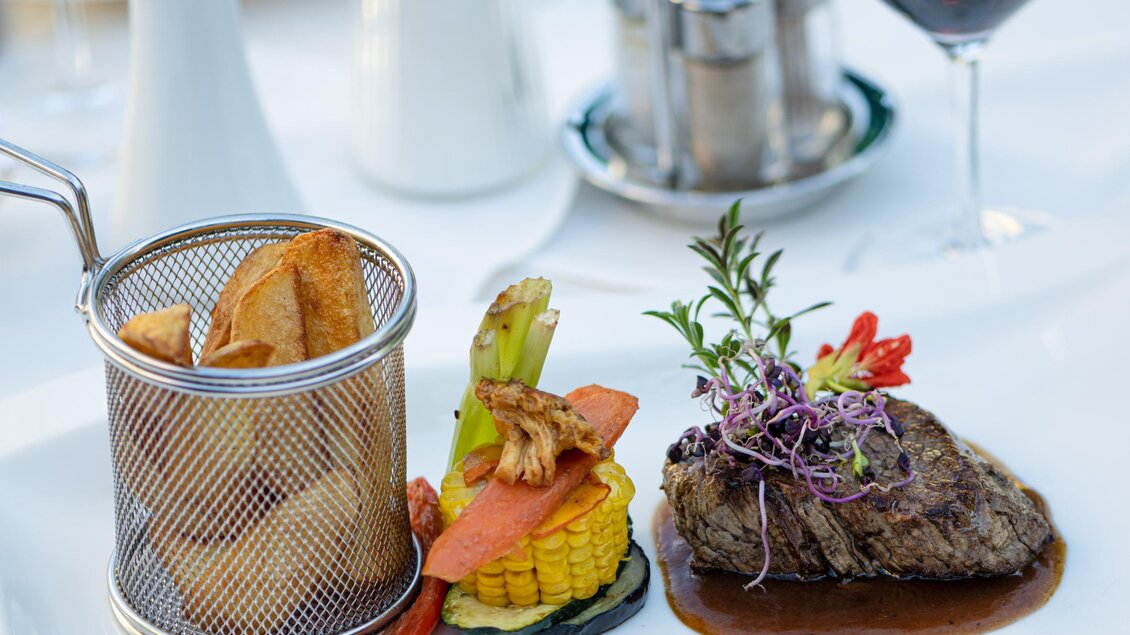 Ein elegantes Gericht auf einem weißen Teller, bestehend aus Fleisch, Gemüse und einer Portion Pommes frites in einem kleinen Korb. Daneben steht ein Glas Rotwein. | © Martina Siebenhandl