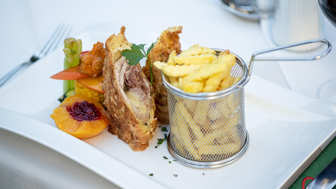 Ein schön angerichteter Teller mit paniertem Fleisch, serviert mit Pommes frites in einem kleinen Metallkorb. Dazu sind bunte Gemüsestücke und Fruchtstücke dekoriert. | © Martina Siebenhandl