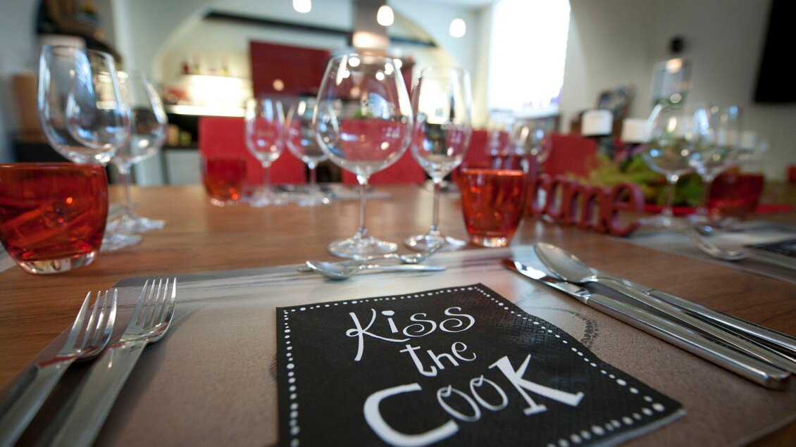 Ein schön gedeckter Tisch mit Glaswaren und Besteck. Auf der Tischdecke steht "Kiss the Cook". | © Kitchen12 - Christian Jungwirth