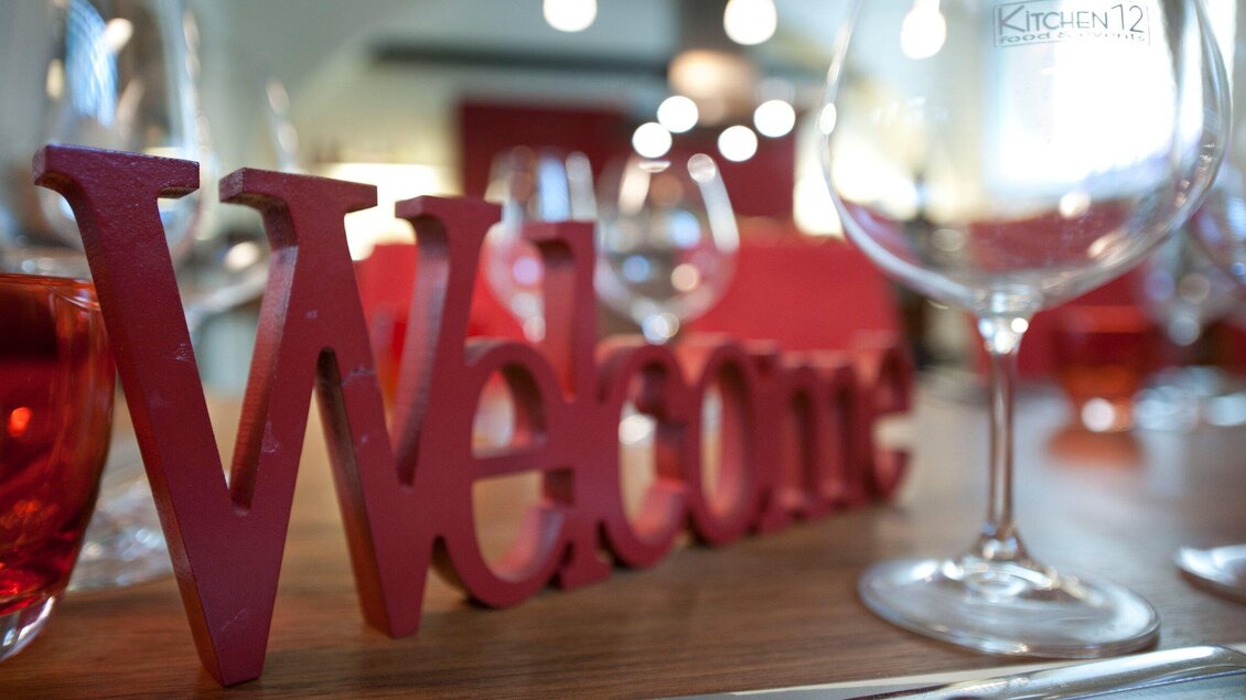 Ein rotes "Welcome"-Schild auf einem Tisch mit Gläsern im Hintergrund. Die Atmosphäre ist einladend und gemütlich. | © Kitchen12 - Christian Jungwirth