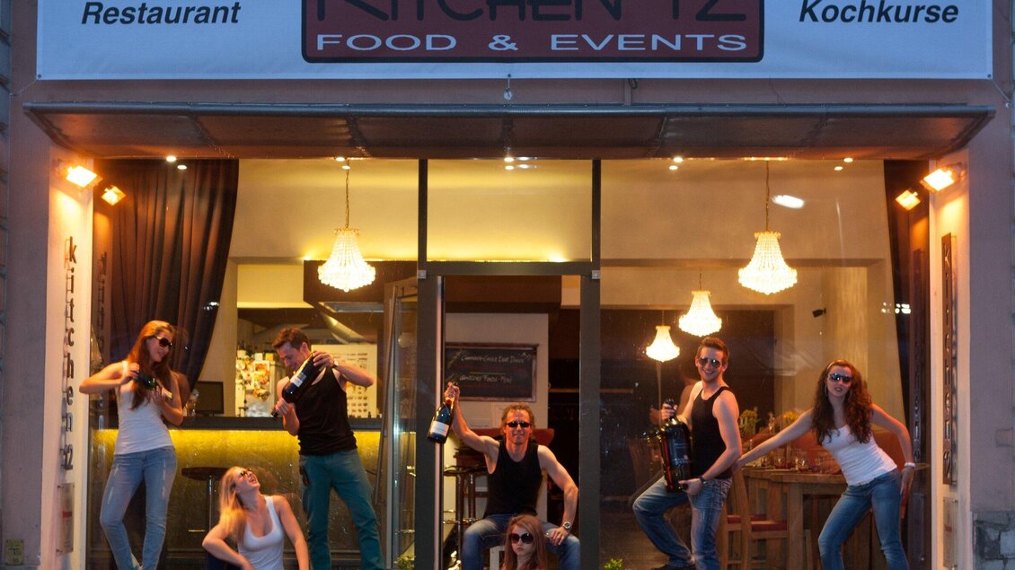 Eine Gruppe von Menschen steht vor einem Restaurant mit dem Namen "Kitchen 12". Sie posieren fröhlich und halten Flaschen in der Hand. | © Kitchen12