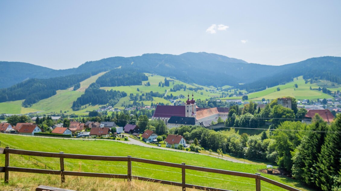Eine Holzbank mit Blick auf eine grüne Landschaft und sanfte Hügel. Im Hintergrund sind ein kleiner Ort und blauer Himmel zu sehen. | © TVB Murau