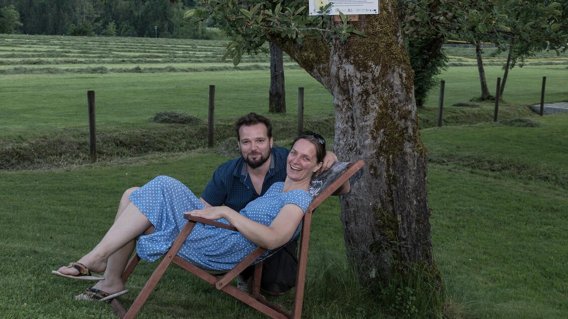 Ein Paar sitzt entspannt in einer Liege unter einem Baum. Im Hintergrund sind grüne Wiesen und ein Zaun zu sehen.