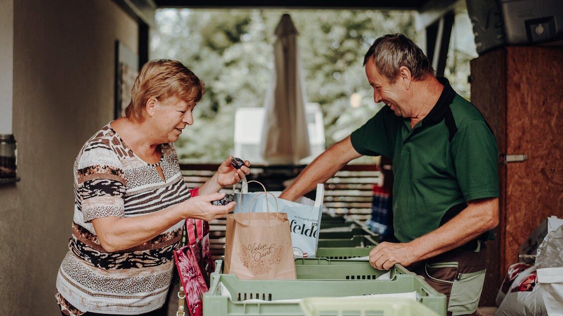 Eine Frau und ein Mann stehen an einem Tisch und tauschen Einkaufstaschen aus. Im Hintergrund sind grüne Kisten und eine gemütliche Umgebung sichtbar. | © GenussReich
