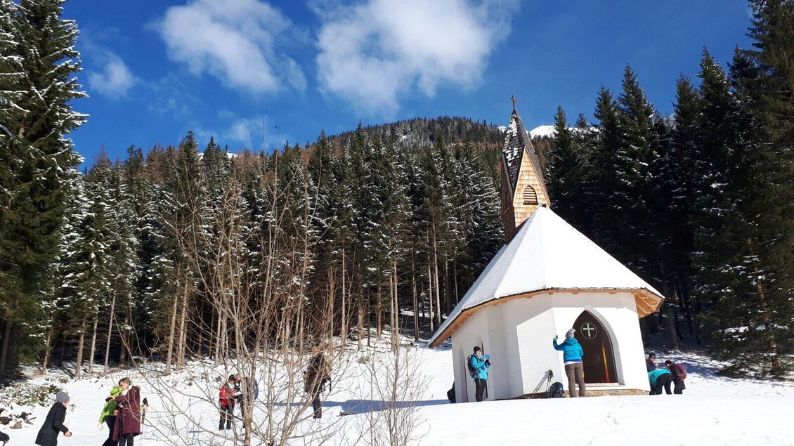 Eine kleine Kapelle im Schnee, umgeben von hohen Tannenbaum. Einige Menschen genießen die winterliche Landschaft. | © Erlebnisregion Murtal