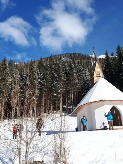 A small chapel in the snow, surrounded by tall fir trees. Some people enjoy the winter landscape. | © Erlebnisregion Murtal
