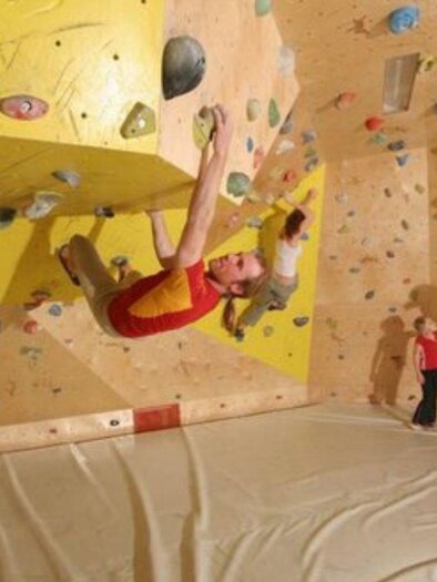 A climbing wall with colorful holds in a climbing gym. A climber hangs on the wall while two spectators watch. | © Mirja Geh