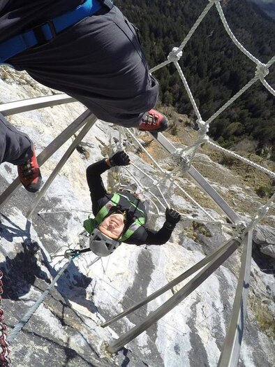 A climber swings on a safety net in the mountains. In the background, forests and a majestic landscape can be seen. | © Martin Hörzer