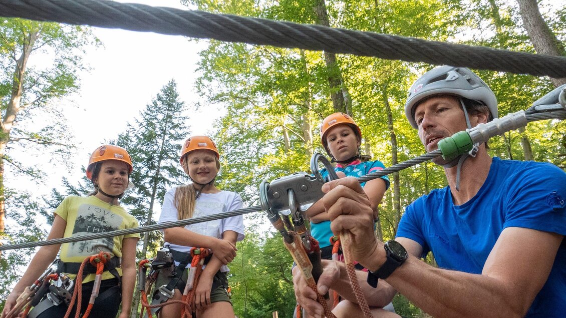 Eine Gruppe von Kindern in Schutzhelmen beobachtet einen Trainer, der ihnen das Sichern am Seil erklärt. Sie stehen in einem Wald umgeben von Bäumen. | © WAP