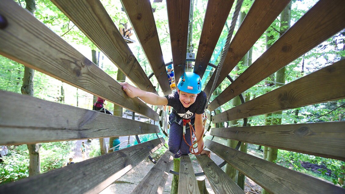 Ein junger Kletterer in einem Abenteuerpark bewegt sich durch eine Holzstruktur. Er trägt einen Helm und lächelt fröhlich. | © WAP