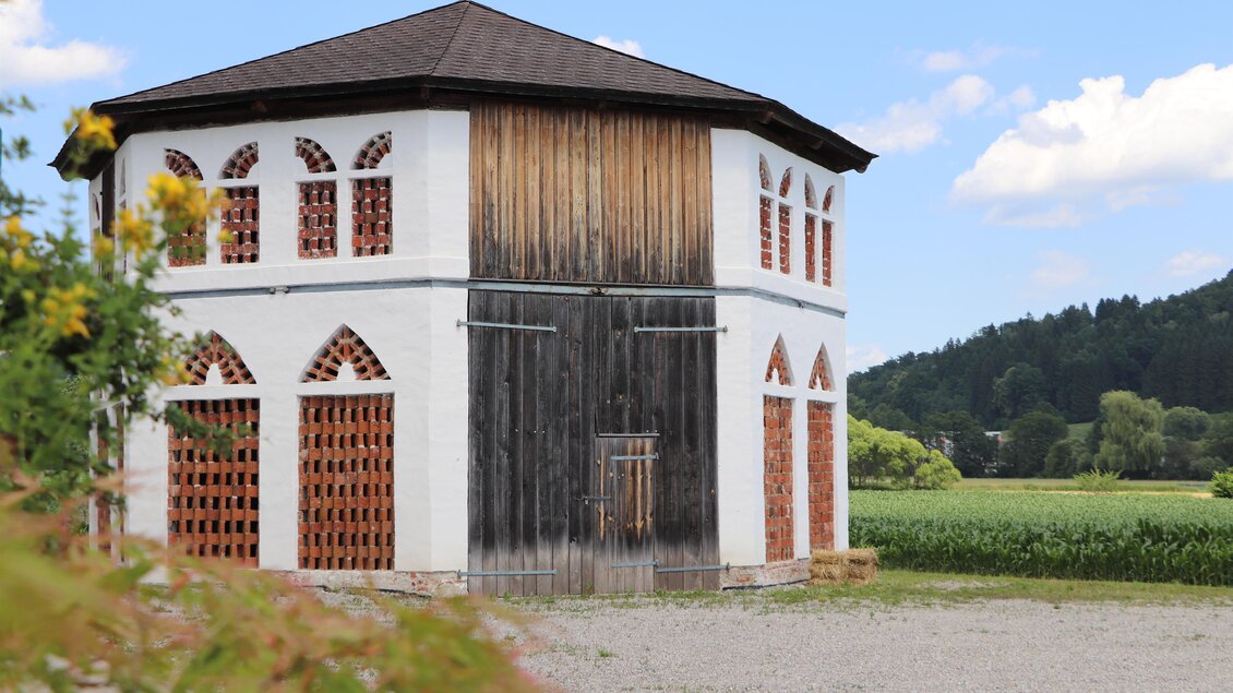 Ein einzigartiges, architektonisch interessantes Gebäude mit Holzelementen und bunten Fenstern. Im Hintergrund erstreckt sich eine grüne Landschaft unter einem klaren, blauen Himmel. | © Marktgemeinde Vasoldsberg