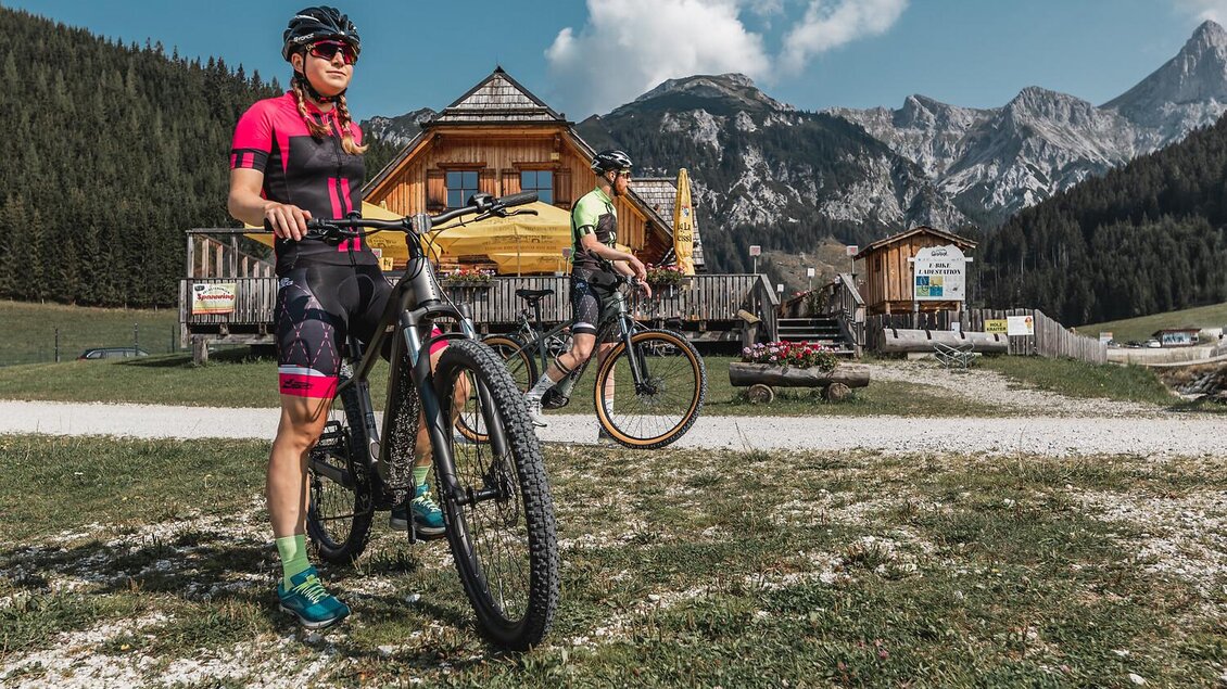 Zwei Radfahrer stehen in einer bergigen Landschaft, umgeben von Natur. Im Hintergrund ist eine gemütliche Berghütte zu sehen.