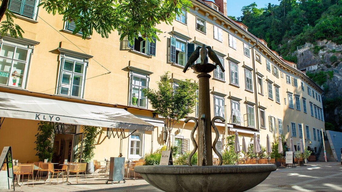 Ein charmantes Restaurant mit Außenbereich und einem Brunnen im Vordergrund. Im Hintergrund sind sonnige, farbenfrohe Gebäude mit grünen Fensterläden zu sehen. | © Klyo