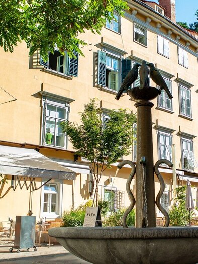 A charming restaurant with an outdoor area and a fountain in the foreground. In the background, sunny, colorful buildings with green shutters can be seen. | © Klyo
