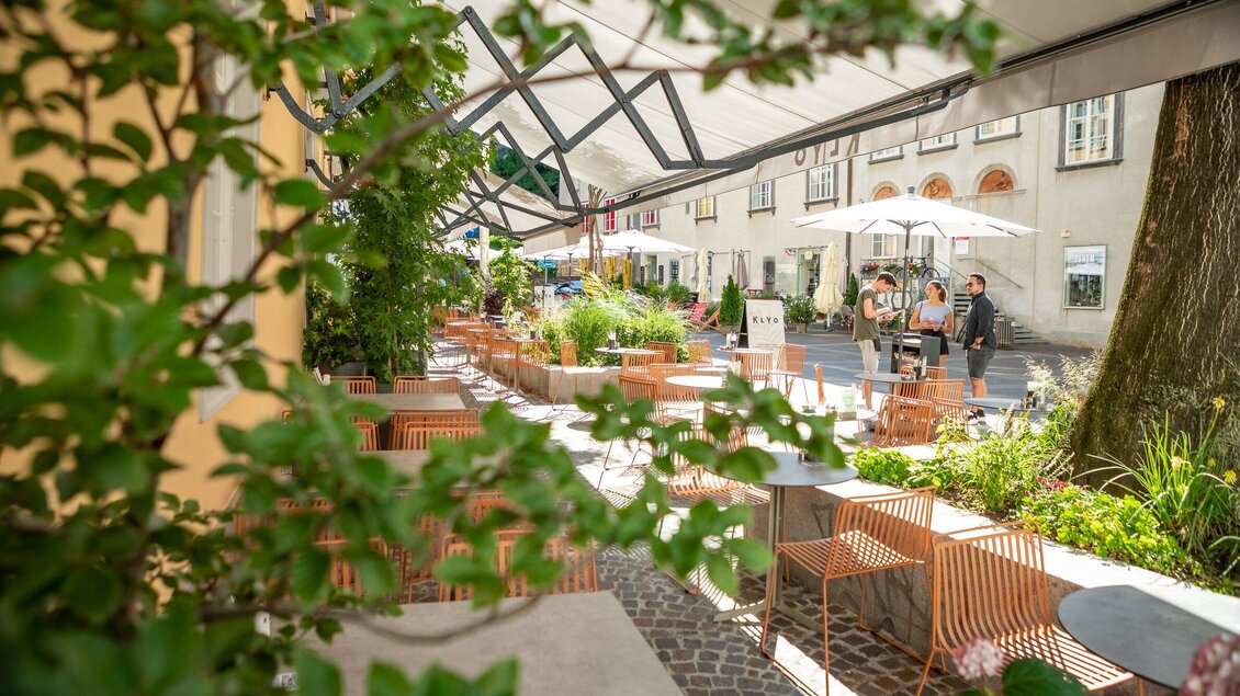 Ein einladendes Café im Freien mit modernen Möbeln und Sonnenschirmen. Üppige Pflanzen umgeben den Bereich, der eine angenehme Atmosphäre schafft. | © Klyo