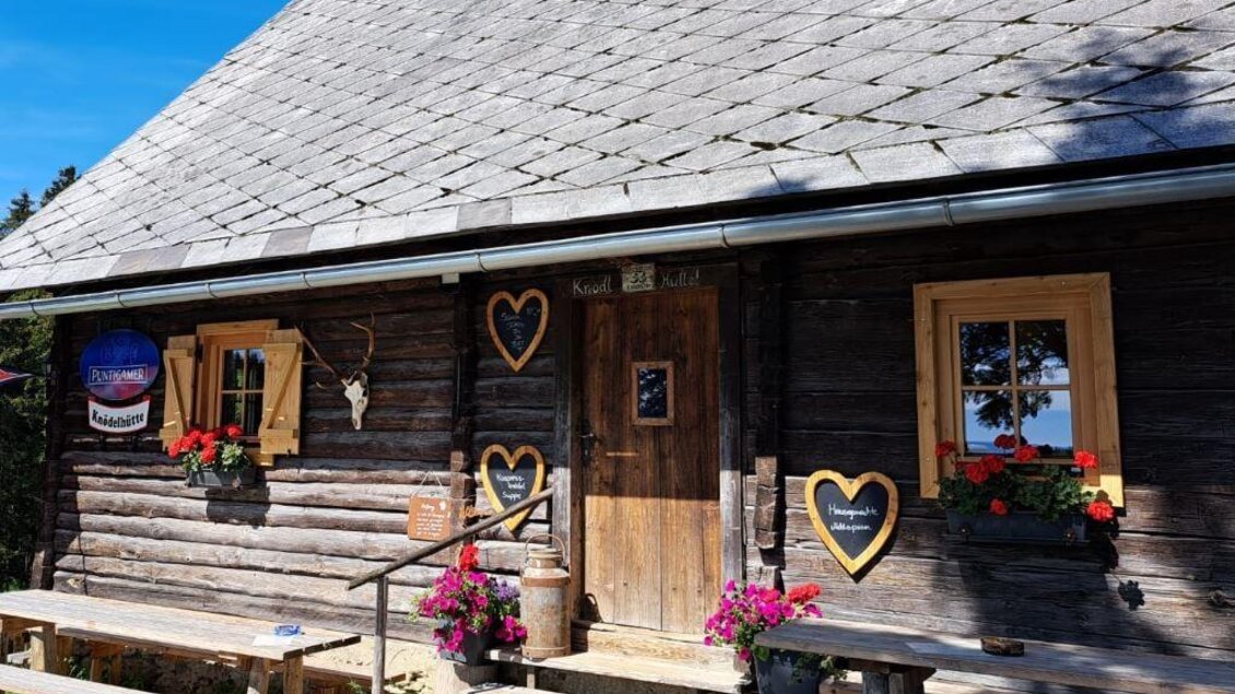 <p>Holzfassade der Knödelhütte mit Eingangstür, Blumenschmuck und Sitzbänken bei Hirschegg-Pack. </p> | © Mathias Renhart