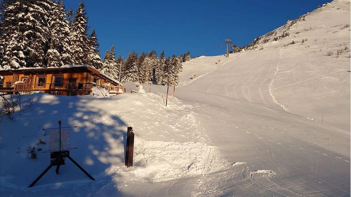 Eine Winterlandschaft mit schneebedeckten Hügeln und einem Holzhaus. Der Himmel ist klar und blau, ideal für Skiausflüge. | © Hirner