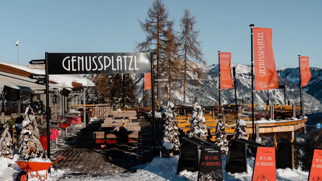 Ein gemütlicher Platz im Schnee namens Genussplatzl mit Blick auf die Berge. Bunte Flaggen und rustikale Möbel schaffen eine einladende Atmosphäre. | © Knapplhütte
