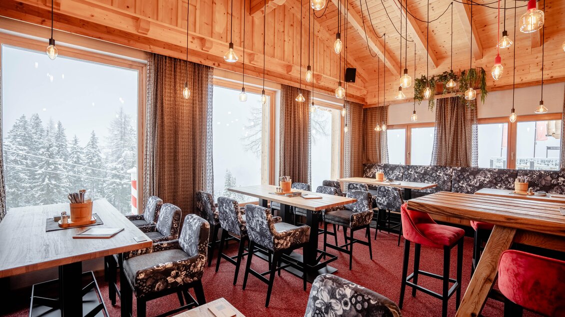 Ein gemütliches Restaurant mit Holzwänden und modernen Sitzplätzen. Durch große Fenster ist eine winterliche Schneelandschaft sichtbar. | © Knapplhütte